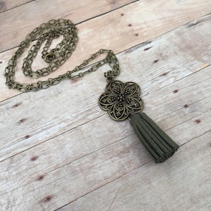 Puede incluir: Un collar de borla de ante verde con un colgante de filigrana de bronce en forma de flor. El collar está en una cadena.