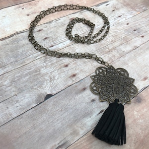 Könnte beinhalten: Eine lange goldene Kettenkette mit einem großen, verzierten, bronzefarbenen Anhänger mit einem Mandala-Design. Der Anhänger ist an einem schwarzen Quasten befestigt.