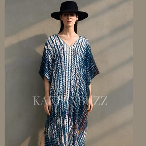 May include: A long, flowing blue and white tie-dye dress with a V-neck and side slits. The dress is paired with a black wide-brimmed hat. The text "KAFTANBUZZ" is visible on the dress.
