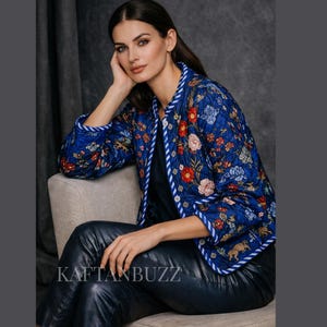 May include: A quilted blue jacket featuring a vibrant floral and animal print in shades of red, white, and gold. The jacket has a round neckline, long sleeves, and a white and blue striped trim along the front opening and cuffs. The text KAFTANBUZZ is visible.