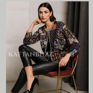 May include: A black jacket with a colorful floral patchwork design. The jacket has long sleeves and a round neckline. The text "KAFTANBUZZ" is visible on the left side of the image. The model is wearing black leather pants and black high heels.