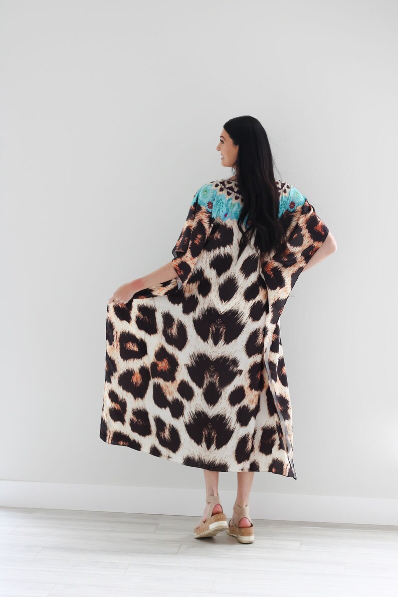 K&ouml;nnte beinhalten: Eine lange, flie&szlig;ende Kaftan mit Leopardenmuster und einem blauen und wei&szlig;en Blumendesign am Ausschnitt. Die Kaftan wird von einer Person mit langen schwarzen Haaren getragen und von hinten gezeigt.