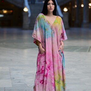 Może przedstawiać: Długa, zwiewna sukienka z dekoltem w serek i wzorem tie-dye w odcieniach r&oacute;żu, błękitu i ż&oacute;łci. Sukienka ma kieszenie i kr&oacute;tkie rękawy. Modelka stoi w dużej, otwartej przestrzeni z marmurową podłogą.