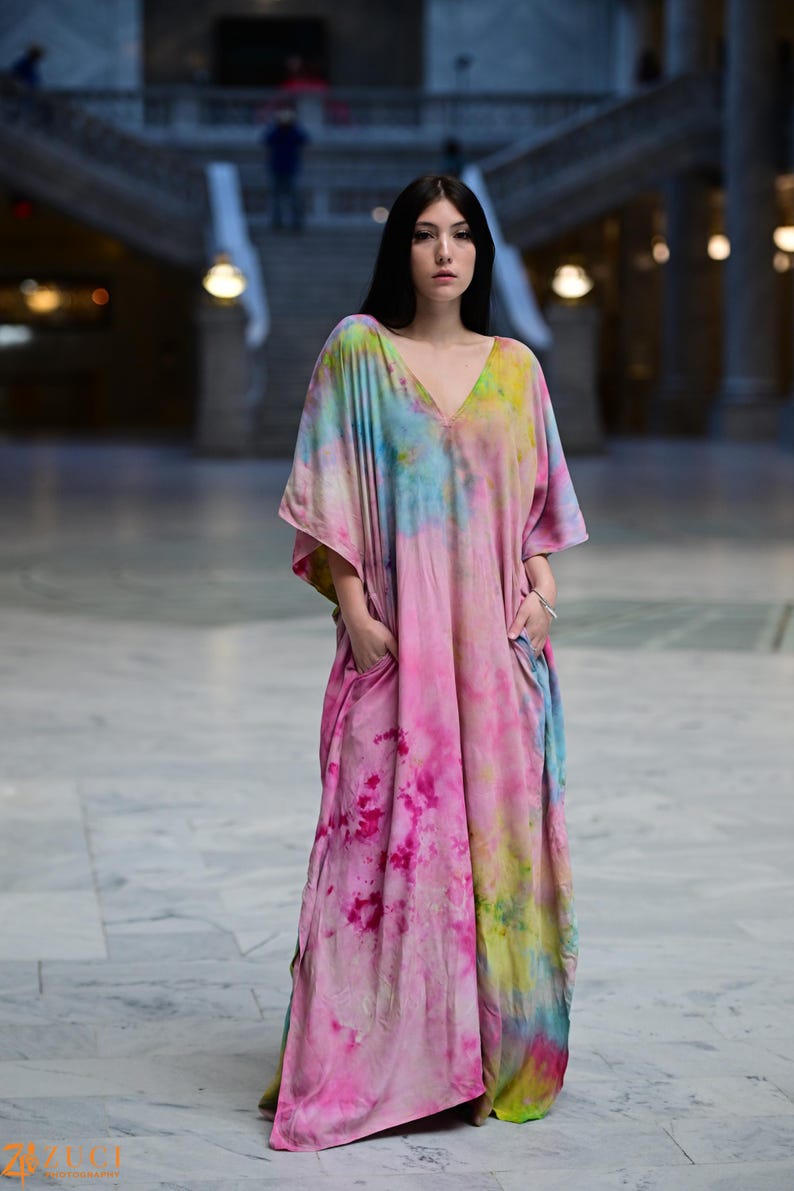 Może przedstawiać: Długa, zwiewna sukienka z dekoltem w serek i wzorem tie-dye w odcieniach r&oacute;żu, błękitu i ż&oacute;łci. Sukienka ma kieszenie i kr&oacute;tkie rękawy. Modelka stoi w dużej, otwartej przestrzeni z marmurową podłogą.