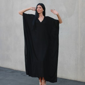 May include: A woman wearing a black maxi dress with a V-neck and long sleeves. She is standing in front of a gray wall and is wearing sandals.