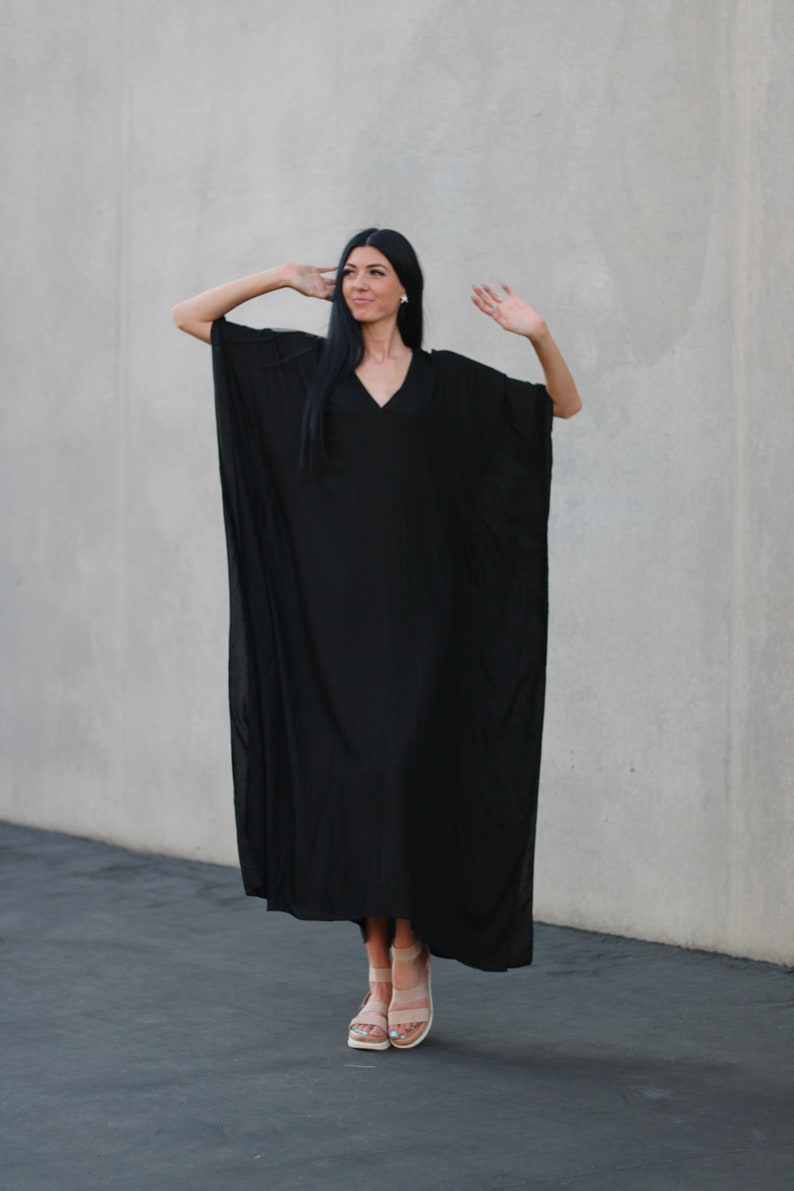 May include: A woman wearing a black maxi dress with a V-neck and long sleeves. She is standing in front of a gray wall and is wearing sandals.