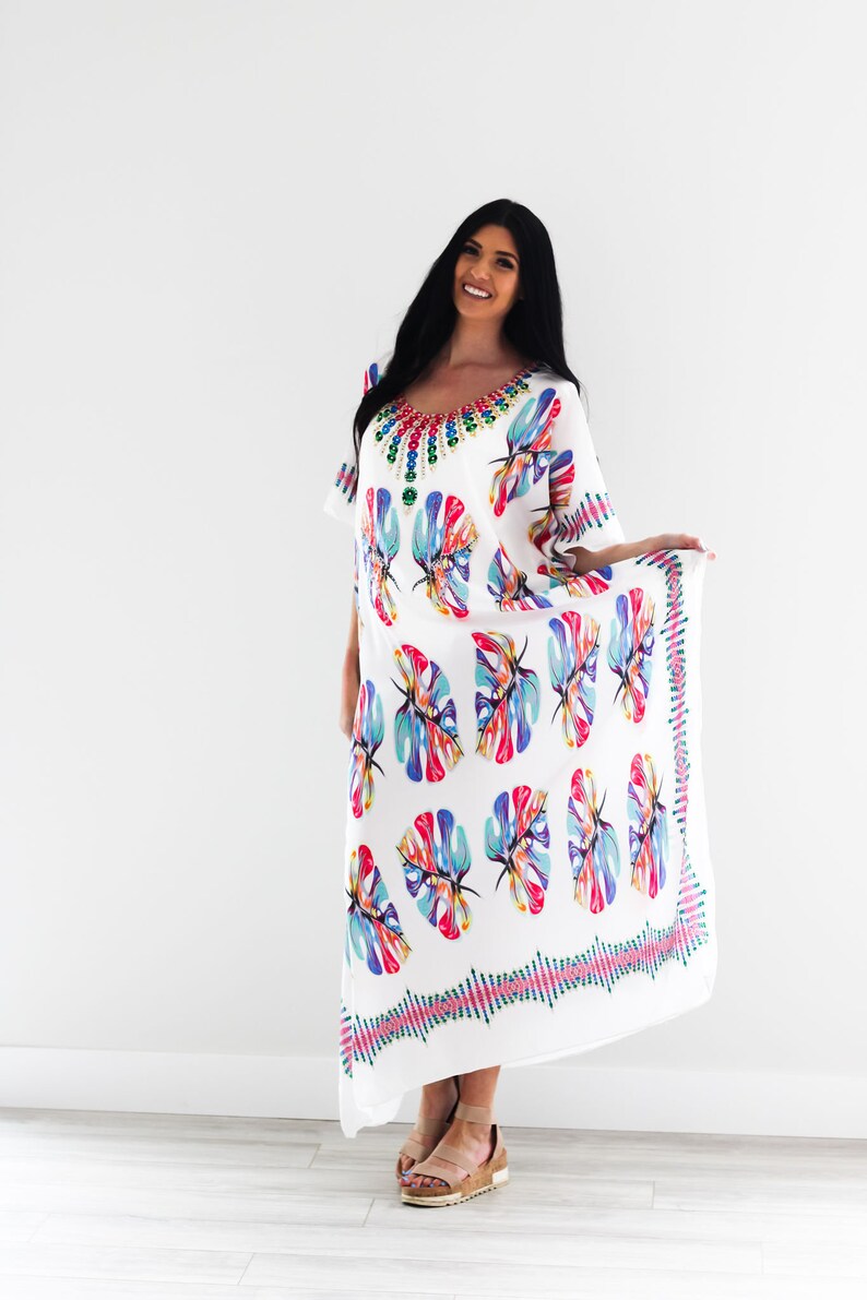 May include: White maxi dress with a colourful floral print and a jeweled neckline. The dress has a flowing silhouette and a relaxed fit.