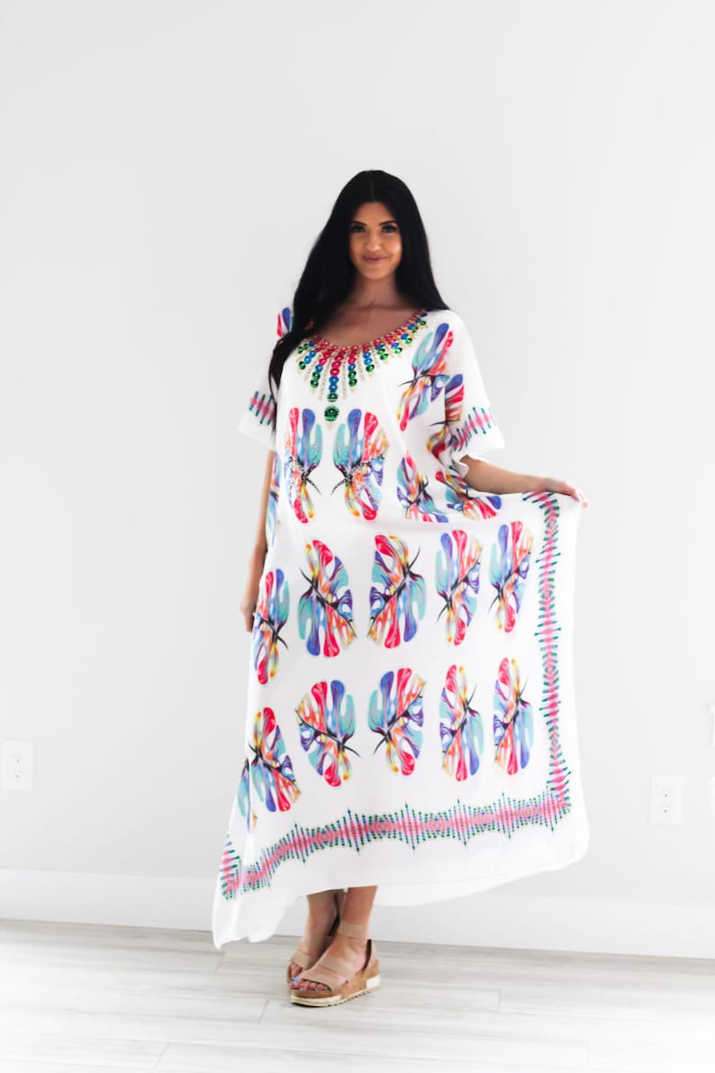 May include: White maxi dress with a colourful leaf print and a beaded necklace. The dress has a short sleeve and a flowing silhouette.