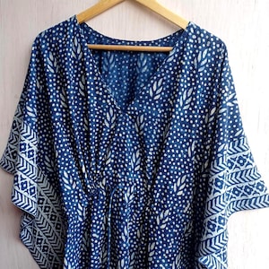 Op de afbeelding: Een blauw en wit patroon kaftan jurk met een V-hals en een taille met trekkoord. De stof heeft een herhalend patroon van witte stippen en bladvormen op een donkerblauwe achtergrond. De mouwen hebben een decoratieve geometrische rand.