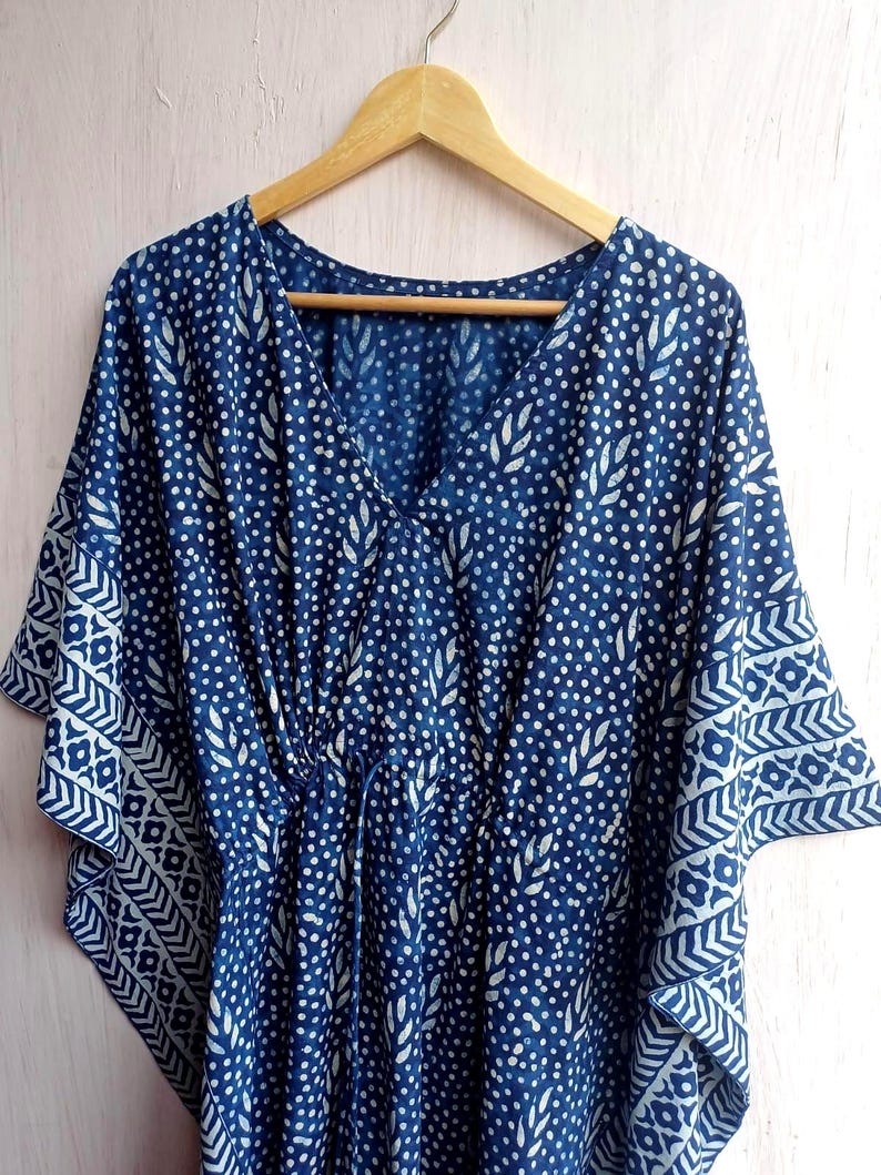 Op de afbeelding: Een blauw en wit patroon kaftan jurk met een V-hals en een taille met trekkoord. De stof heeft een herhalend patroon van witte stippen en bladvormen op een donkerblauwe achtergrond. De mouwen hebben een decoratieve geometrische rand.