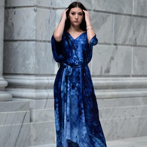 May include: A long, flowing blue tie-dye dress with a V-neck and a matching belt. The dress has a loose, comfortable fit and is paired with black high-heeled sandals. The model is standing in front of a marble wall.