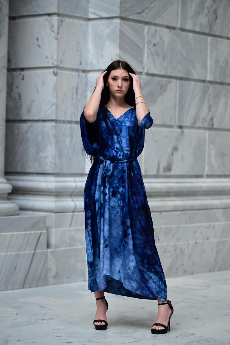 May include: A long, flowing blue tie-dye dress with a V-neck and a matching belt. The dress has a loose, comfortable fit and is paired with black high-heeled sandals. The model is standing in front of a marble wall.