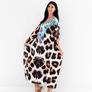 K&ouml;nnte beinhalten: Eine Frau tr&auml;gt eine lange, flie&szlig;ende Kaftan mit Leopardenmuster und einem t&uuml;rkisfarbenen und floralen Design am Oberteil. Sie tr&auml;gt Sandalen und l&auml;chelt in die Kamera.