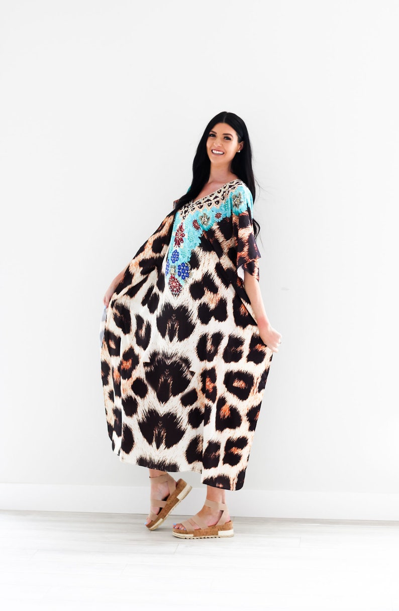 K&ouml;nnte beinhalten: Eine Frau tr&auml;gt eine lange, flie&szlig;ende Kaftan mit Leopardenmuster und einem t&uuml;rkisfarbenen und floralen Design am Oberteil. Sie tr&auml;gt Sandalen und l&auml;chelt in die Kamera.