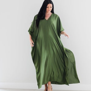 May include: A woman wearing a long, flowing, olive green kaftan dress with a V-neckline. She is standing on a light wood floor and wearing tan sandals.