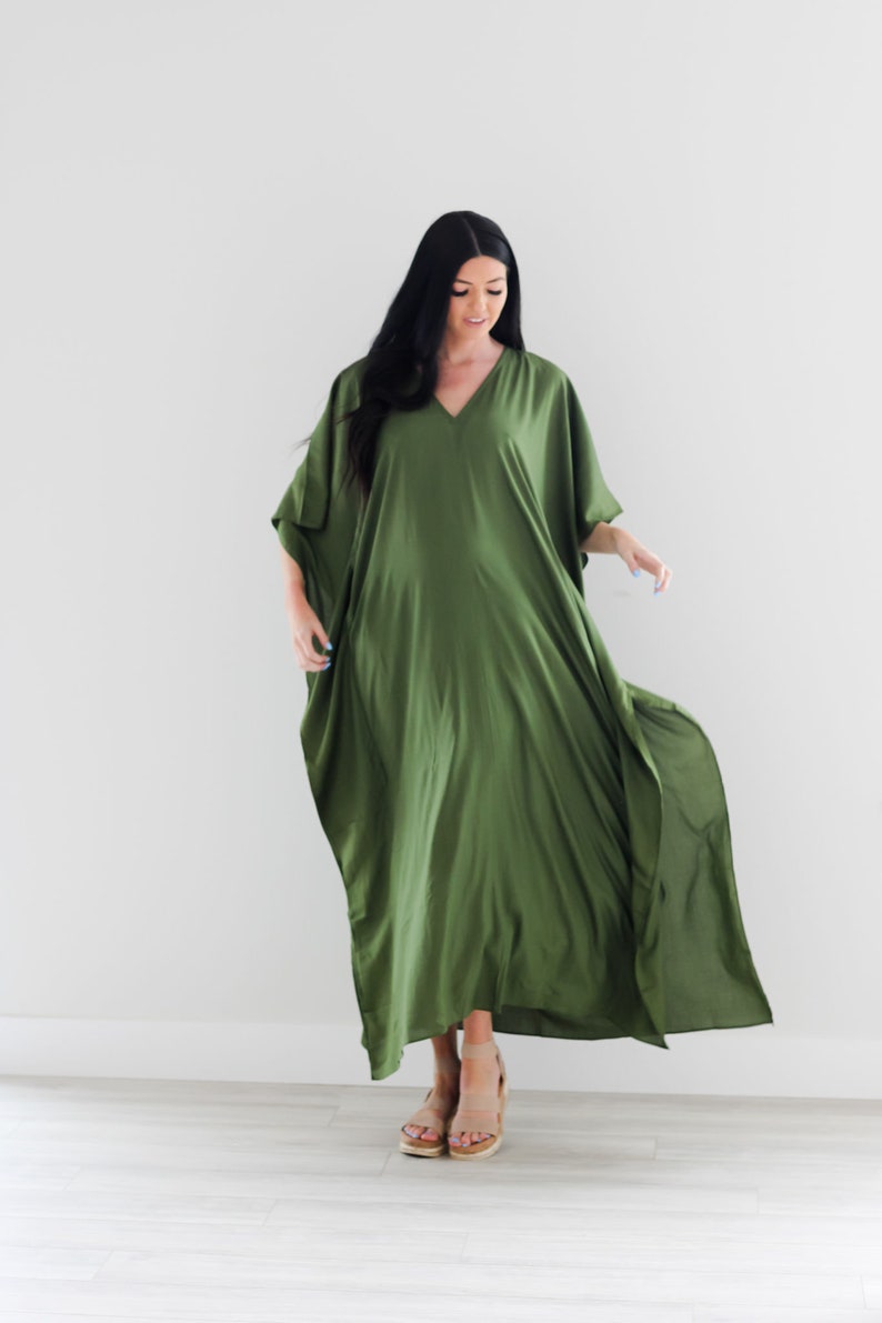 May include: A woman wearing a long, flowing, olive green kaftan dress with a V-neckline. She is standing on a light wood floor and wearing tan sandals.