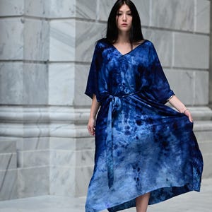 May include: A flowing, tie-dye blue dress with a V-neck and a matching belt. The dress has short sleeves and a long, asymmetrical hemline. The model is wearing black high-heeled sandals. The background is a marble wall.