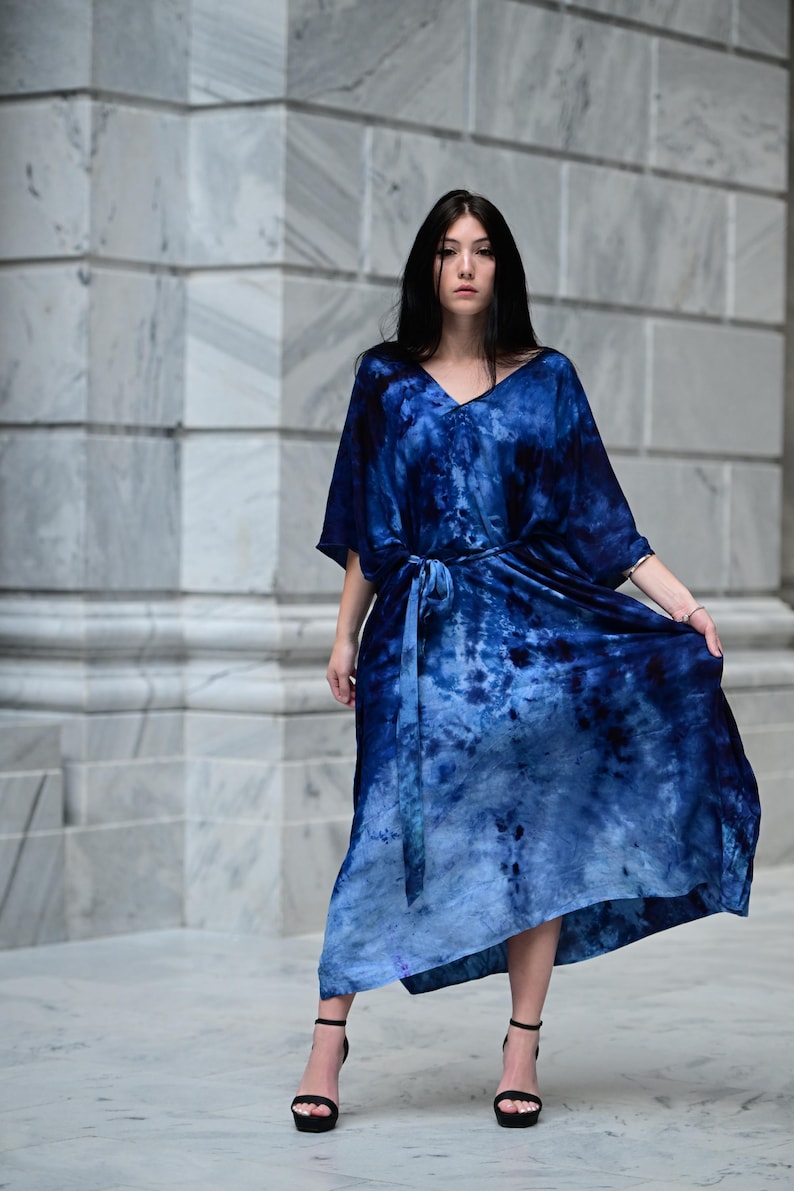 May include: A flowing, tie-dye blue dress with a V-neck and a matching belt. The dress has short sleeves and a long, asymmetrical hemline. The model is wearing black high-heeled sandals. The background is a marble wall.