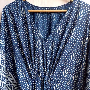 Op de afbeelding: Een marineblauwe kaftan met een V-hals en een taille met trekkoord. De stof is bedrukt met witte stippen en bladvormige patronen. De kaftan heeft een decoratieve rand met geometrische ontwerpen.