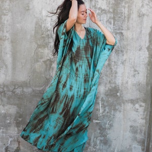 Pode incluir: Uma mulher vestindo um longo vestido kaftan de cor turquesa e marrom com estampa tie-dye. O vestido tem um decote em V e mangas compridas. Ela est&aacute; em p&eacute; em frente a uma parede de concreto. O texto "KAFTAN BUZZ" &eacute; vis&iacute;vel no vestido.