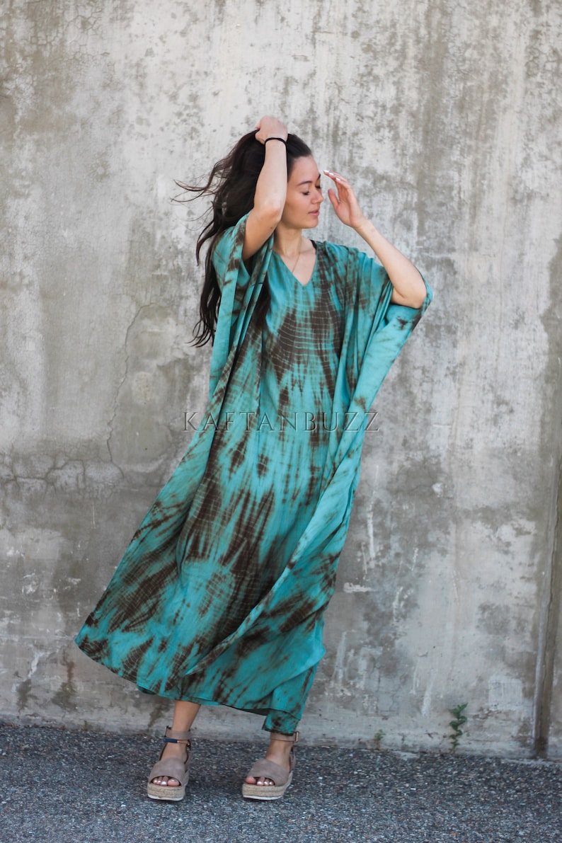 Pode incluir: Uma mulher vestindo um longo vestido kaftan de cor turquesa e marrom com estampa tie-dye. O vestido tem um decote em V e mangas compridas. Ela est&aacute; em p&eacute; em frente a uma parede de concreto. O texto "KAFTAN BUZZ" &eacute; vis&iacute;vel no vestido.