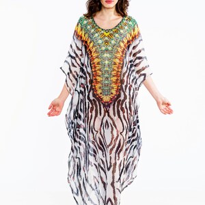 May include: A flowing, long caftan dress with a V-neckline. The dress features a vibrant, patterned yoke in shades of yellow, orange, and green. The body of the dress has a zebra stripe pattern in black and white. The model is wearing beige platform sandals.