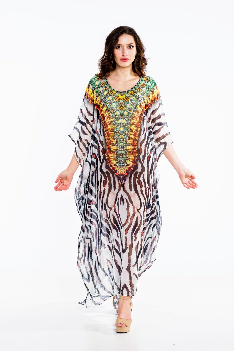 May include: A flowing, long caftan dress with a V-neckline. The dress features a vibrant, patterned yoke in shades of yellow, orange, and green. The body of the dress has a zebra stripe pattern in black and white. The model is wearing beige platform sandals.