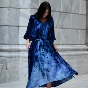 May include: A long, flowing blue dress with a tie-dye pattern. The dress features a V-neck, wide sleeves, and a matching belt. The model is wearing black high heels. The dress is a vibrant shade of blue, with lighter shades towards the bottom.