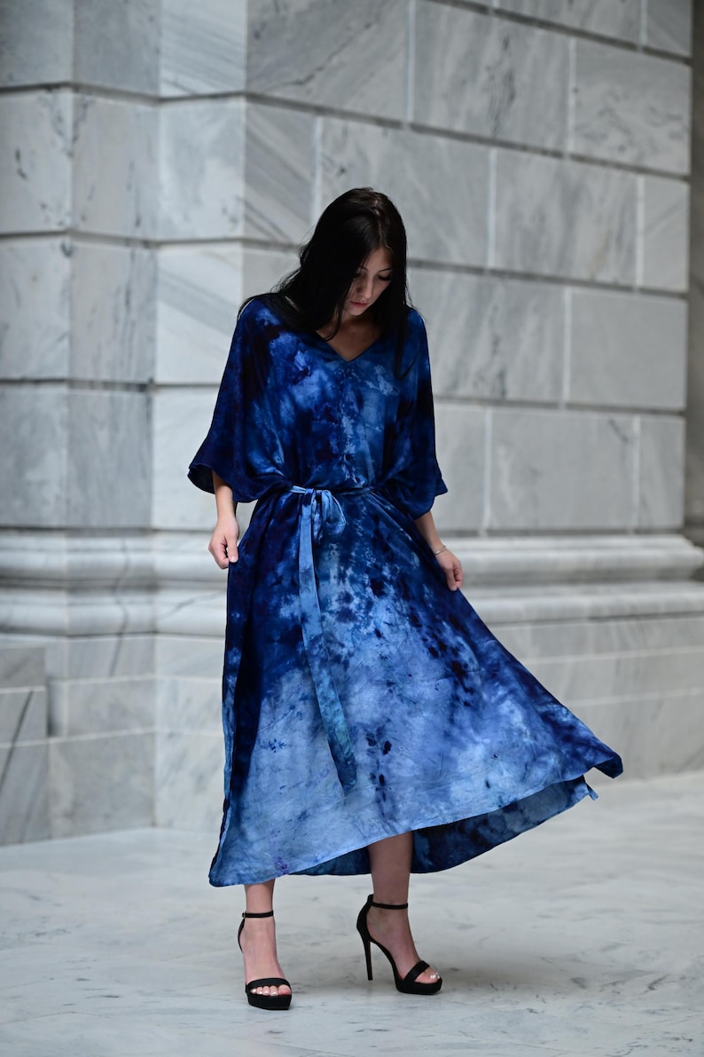 May include: A long, flowing blue dress with a tie-dye pattern. The dress features a V-neck, wide sleeves, and a matching belt. The model is wearing black high heels. The dress is a vibrant shade of blue, with lighter shades towards the bottom.