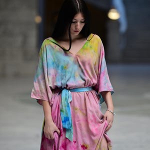 Może przedstawiać: Sukienka maxi w stylu tie-dye z dekoltem w serek i kr&oacute;tkimi rękawami. Sukienka ma r&oacute;żową, niebieską i ż&oacute;łtą kolorystykę, z niebieskim pasem wiązanym w talii. Sukienka jest wykonana z miękkiej, zwiewnej tkaniny.