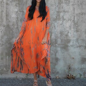 Puede incluir: Una mujer lleva un vestido maxi largo de color naranja y azul con estampado tie-dye, cuello en V y mangas largas. Est&aacute; de pie frente a una pared de hormig&oacute;n y sonr&iacute;e. El vestido tiene un corte fluido y relajado.
