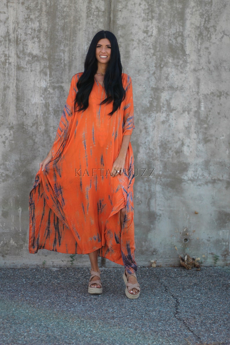 Puede incluir: Una mujer lleva un vestido maxi largo de color naranja y azul con estampado tie-dye, cuello en V y mangas largas. Est&aacute; de pie frente a una pared de hormig&oacute;n y sonr&iacute;e. El vestido tiene un corte fluido y relajado.