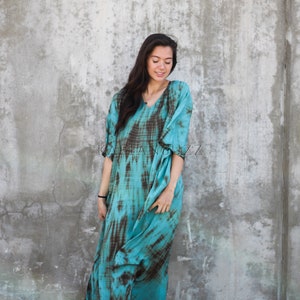 Pode incluir: Uma mulher vestindo um vestido longo e fluido de cor turquesa e marrom com estampa tie-dye e decote em V. Ela est&aacute; em p&eacute; em frente a uma parede de concreto.