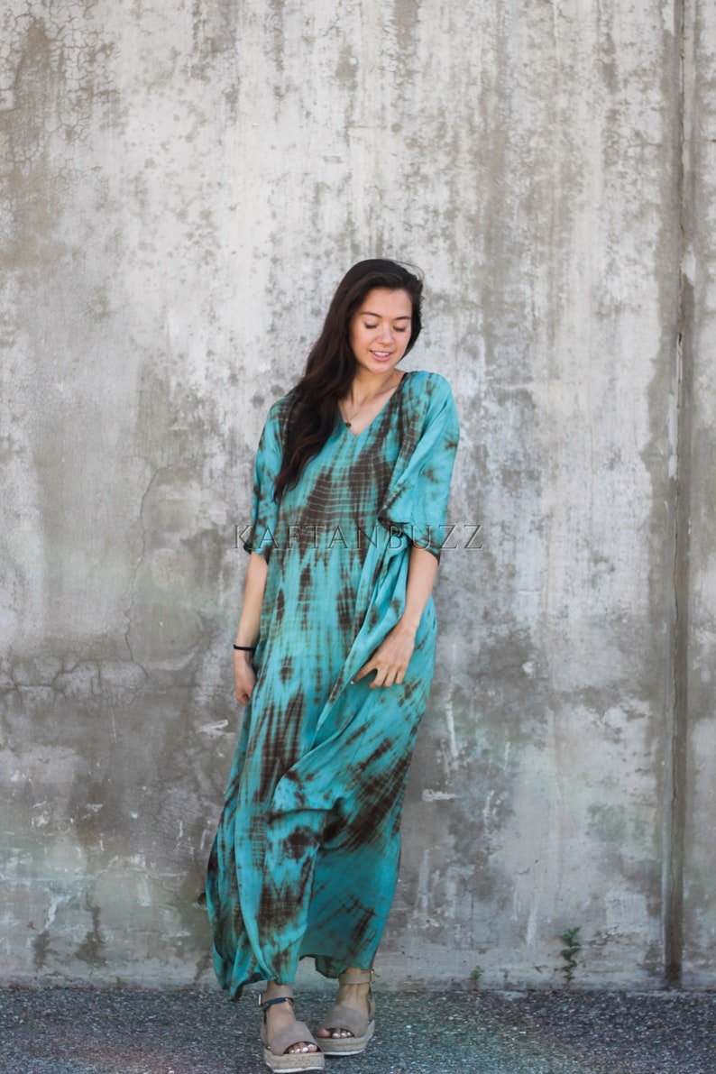 Pode incluir: Uma mulher vestindo um vestido longo e fluido de cor turquesa e marrom com estampa tie-dye e decote em V. Ela est&aacute; em p&eacute; em frente a uma parede de concreto.