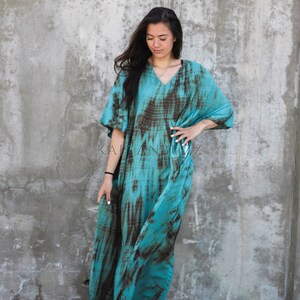 Pode incluir: Uma kaftan turquesa e marrom com estampa tie-dye, decote em V e mangas curtas. A kaftan &eacute; solta e chega aos tornozelos. A modelo est&aacute; usando a kaftan com sand&aacute;lias marrons.