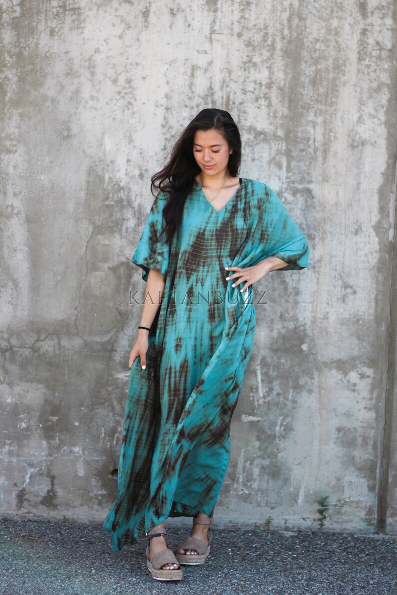 Pode incluir: Uma kaftan turquesa e marrom com estampa tie-dye, decote em V e mangas curtas. A kaftan &eacute; solta e chega aos tornozelos. A modelo est&aacute; usando a kaftan com sand&aacute;lias marrons.