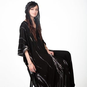 May include: A woman wearing a black and white tie-dye kaftan with long dreadlocks. She is sitting on a black stool and looking at the camera.