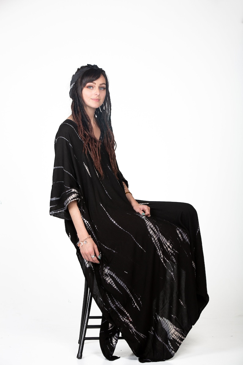 May include: A woman wearing a black and white tie-dye kaftan with long dreadlocks. She is sitting on a black stool and looking at the camera.