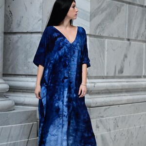 May include: A long, flowing blue tie-dye dress with a deep V-neck and short sleeves. The dress features a gradient of blue hues and is worn with black high-heeled sandals. The model is standing in front of a marble wall.