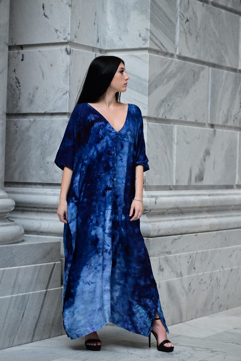 May include: A long, flowing blue tie-dye dress with a deep V-neck and short sleeves. The dress features a gradient of blue hues and is worn with black high-heeled sandals. The model is standing in front of a marble wall.