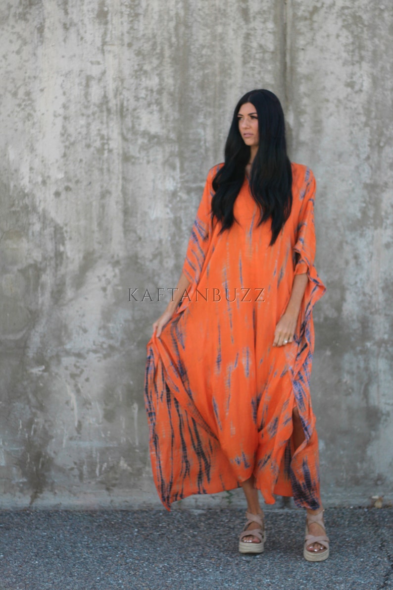 Puede incluir: Una mujer lleva un vestido largo de kaft&aacute;n de color naranja y azul con estampado tie-dye y escote en V. El vestido tiene una silueta fluida y est&aacute; hecho de una tela ligera. La mujer est&aacute; de pie frente a una pared de hormig&oacute;n. El texto "KAFTANBUZZ" es visible en la imagen.