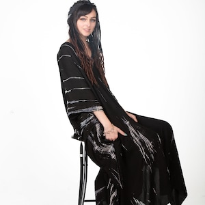 May include: A woman wearing a black and white tie-dye maxi dress with long brown dreadlocks. She is sitting on a black stool and looking at the camera.