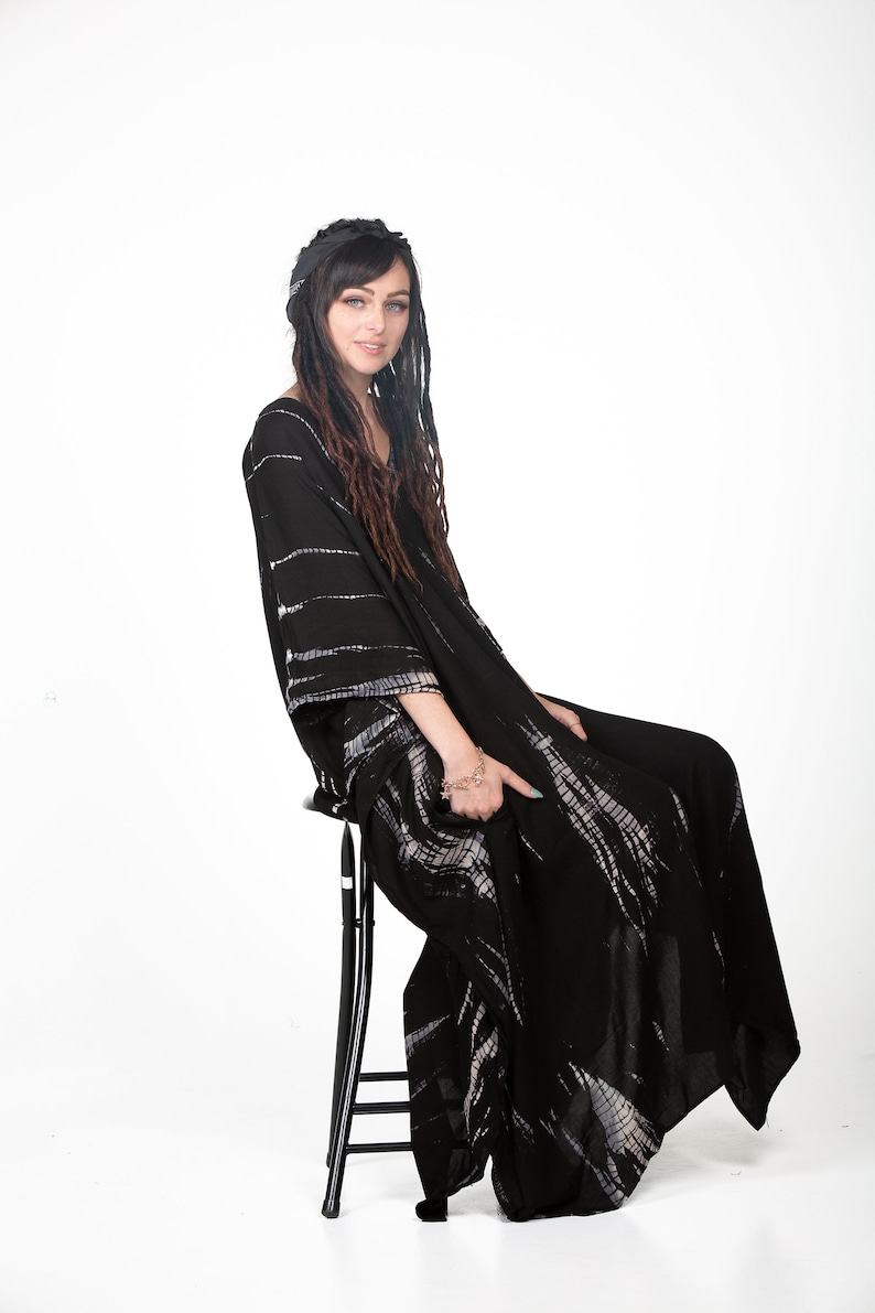 May include: A woman wearing a black and white tie-dye maxi dress with long brown dreadlocks. She is sitting on a black stool and looking at the camera.