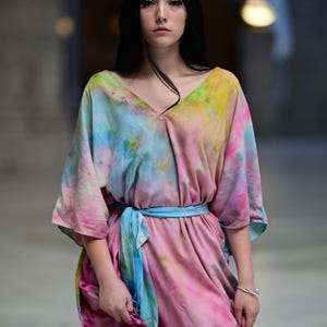 Może przedstawiać: Sukienka typu tie-dye z dekoltem w serek i niebieskim paskiem. Sukienka ma mieszankę r&oacute;żowych, ż&oacute;łtych i niebieskich odcieni. Rękawy są do łokcia. Modelka ma na sobie srebrną bransoletkę. Sukienka to wyjątkowa deklaracja mody.