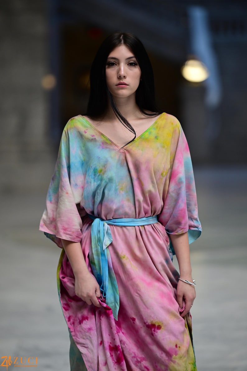 Może przedstawiać: Sukienka typu tie-dye z dekoltem w serek i niebieskim paskiem. Sukienka ma mieszankę r&oacute;żowych, ż&oacute;łtych i niebieskich odcieni. Rękawy są do łokcia. Modelka ma na sobie srebrną bransoletkę. Sukienka to wyjątkowa deklaracja mody.