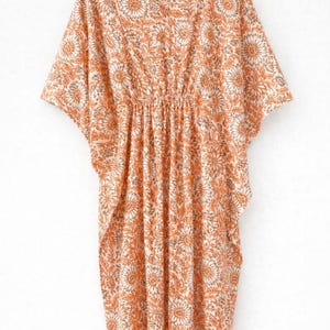 May include: An orange and cream-colored caftan dress with a floral pattern. The dress has short sleeves and a gathered waist. The fabric is lightweight and appears to be cotton or a similar material. The dress is hanging on a wooden hanger.