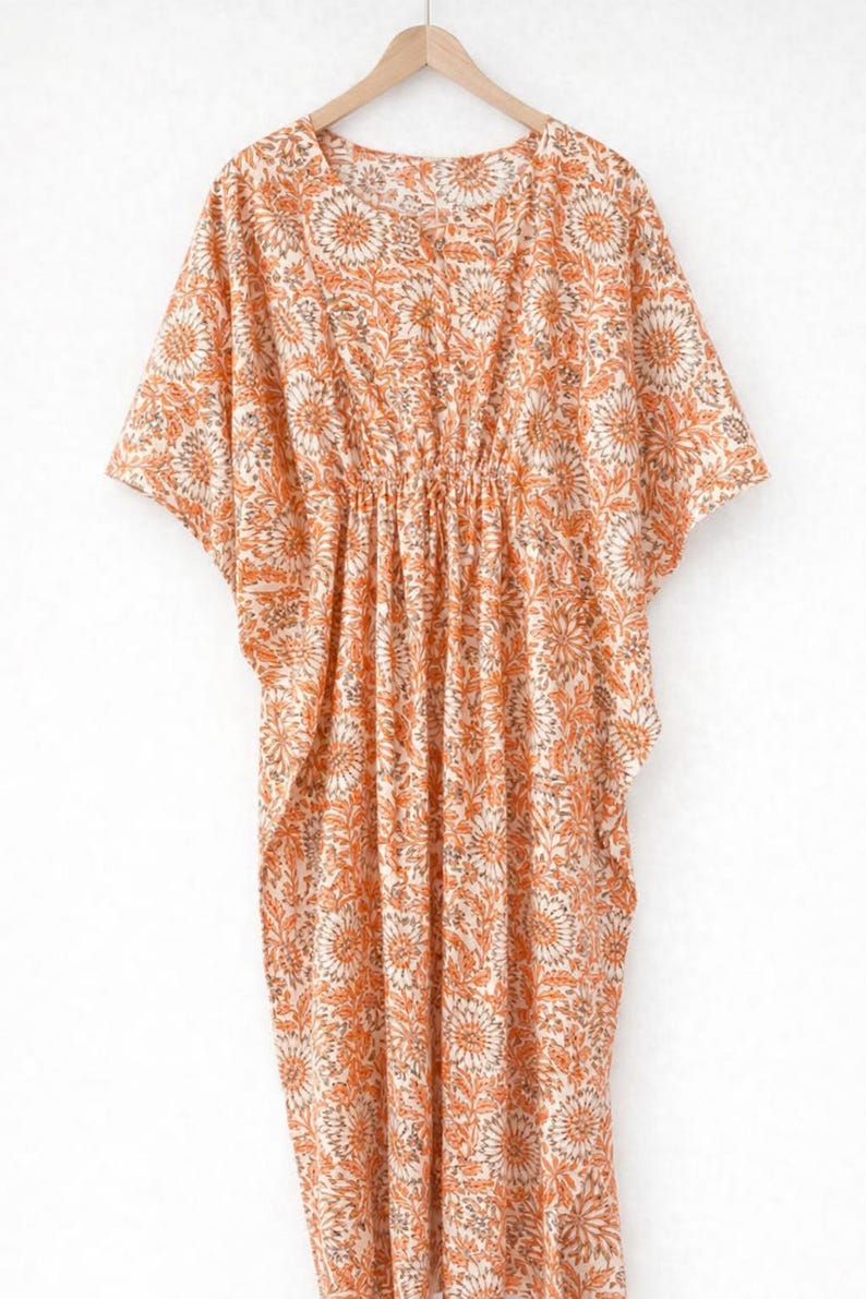May include: An orange and cream-colored caftan dress with a floral pattern. The dress has short sleeves and a gathered waist. The fabric is lightweight and appears to be cotton or a similar material. The dress is hanging on a wooden hanger.