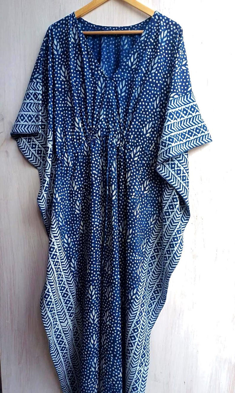 Op de afbeelding: Een blauw en wit patroon caftan jurk. De jurk heeft een V-hals, korte mouwen en een taille met trekkoord. De stof is donkerblauw met een wit stip- en bladerpatroon en witte geometrische ontwerpen op de mouwen.