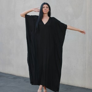 May include: A woman wearing a long, black, flowing dress with a V-neckline. She is standing in front of a gray wall and is wearing sandals.