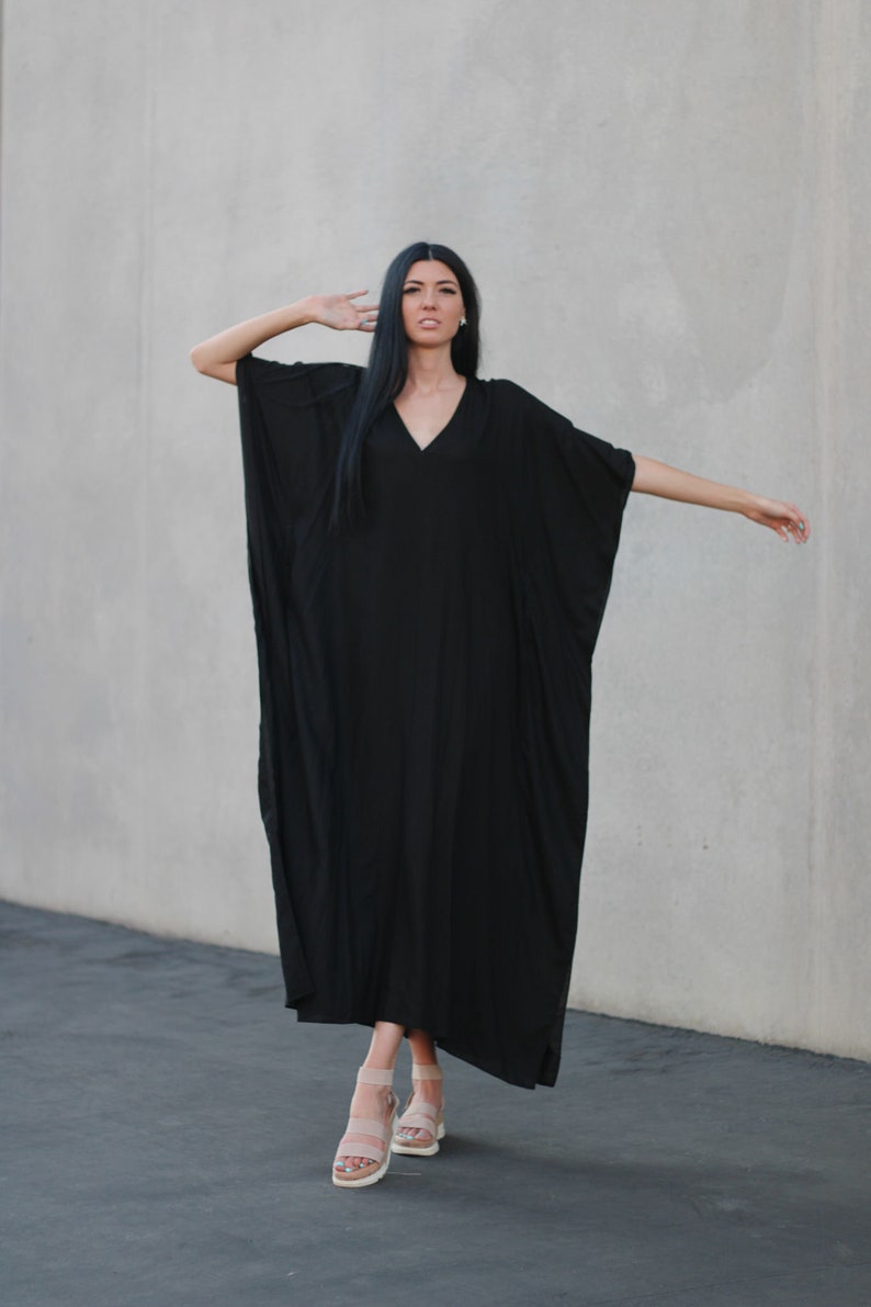 May include: A woman wearing a long, black, flowing dress with a V-neckline. She is standing in front of a gray wall and is wearing sandals.